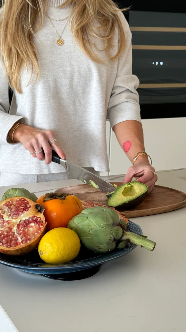 Person cutting an avocado on a wooden cutting board with various fruits and vegetables on a table.